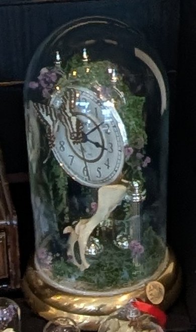 A clock face set inside a glass dome with a small skeleton, moss, and dried flowers on a gold base.