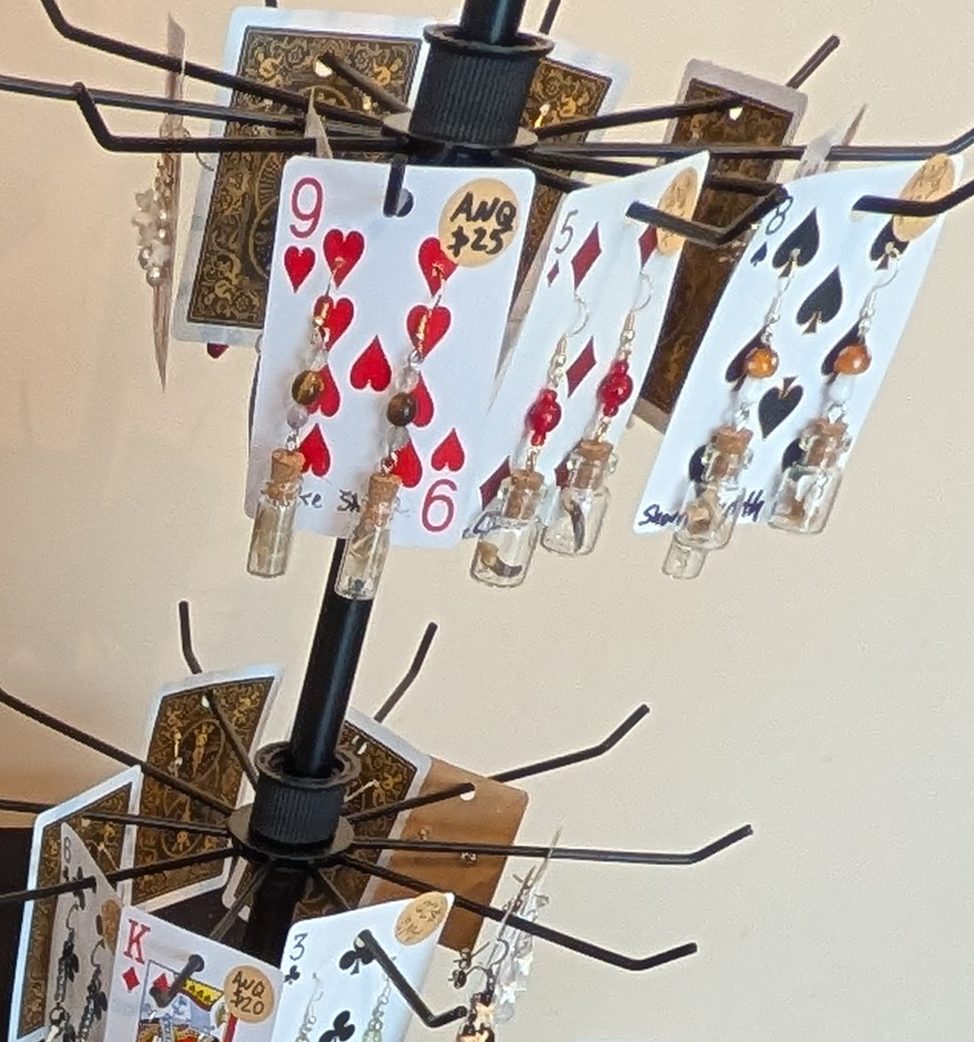 Earrings hung on playing cards on a black spinner rack, including pairs of small cork-stoppered glass vials with beads.