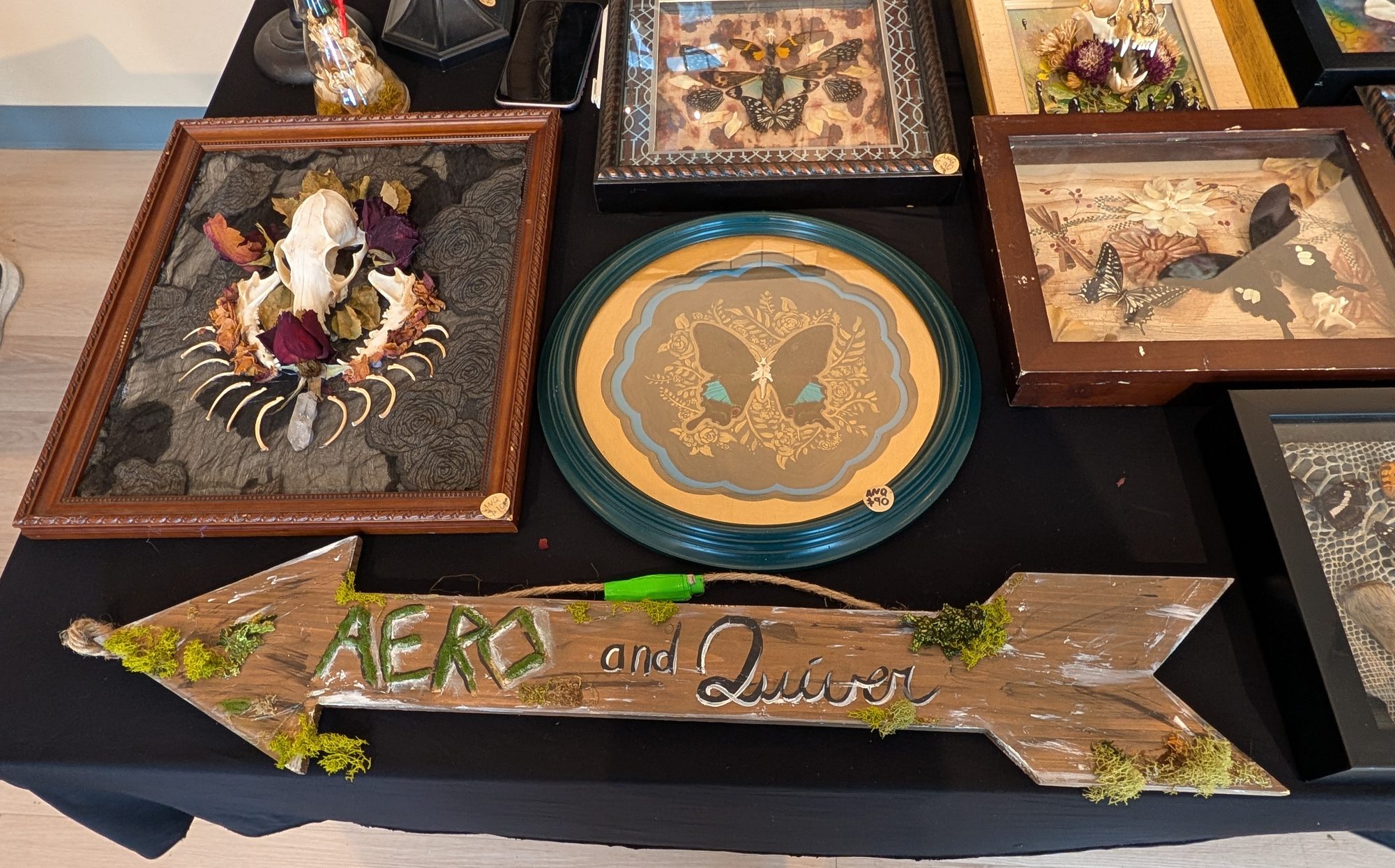 The Aero and Quiver table: a framed skull and claw piece with dried roses, a round teal frame holding a butterfly, framed butterfly collections, and the hand-lettered wooden arrow sign reading Aero and Quiver.